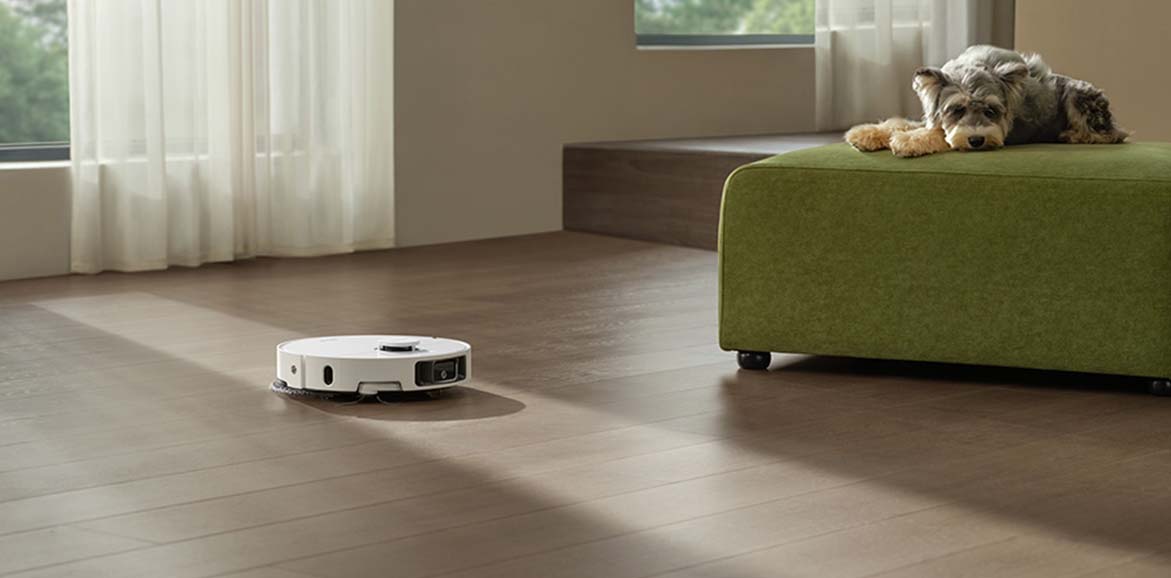 Robot sprzątający porusza się po drewnianej podłodze, obok leży pies na zielonym meblu